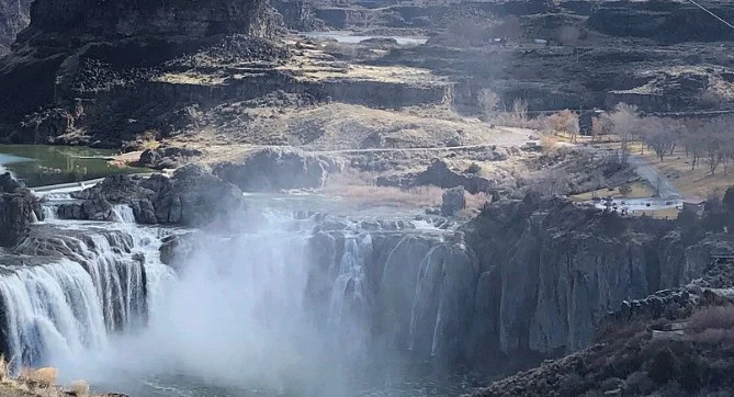 A wide waterfall cascades over rocky cliffs into a river below, surrounded by rugged terrain and sparse vegetation, reminiscent of the breathtaking landscapes found near Twin Falls, Idaho.
