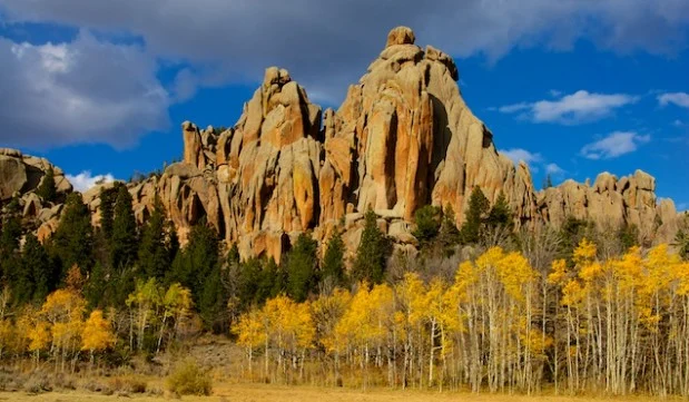 Tall, jagged rock formations rise above a forest of green and yellow trees under a partly cloudy blue sky—an iconic landscape near Laramie, Wyoming.