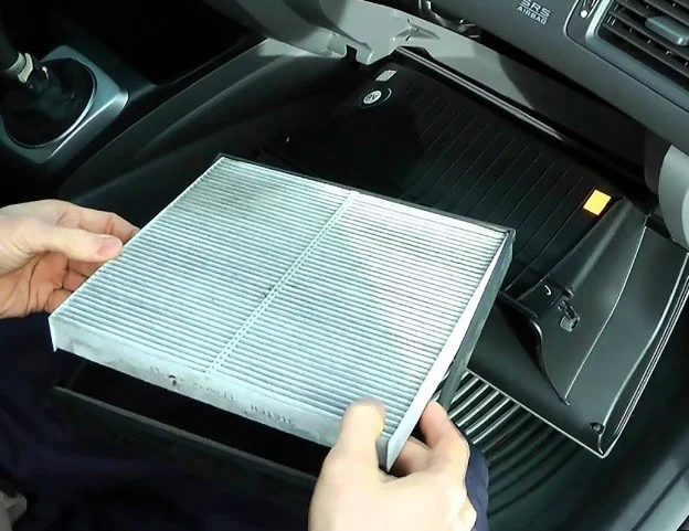 A person holds a rectangular cabin air filter inside a car, with the glove compartment open and the interior dashboard visible—preparing for clean airflow before tackling Yellowstone traffic near Idaho Falls.
