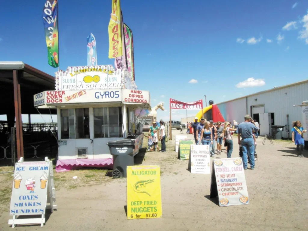 A row of food concession stands at an Elizabeth Ranch fair, with sandwich board signs listing menu items like funnel cakes, deep fried alligator nuggets, and ice cream.