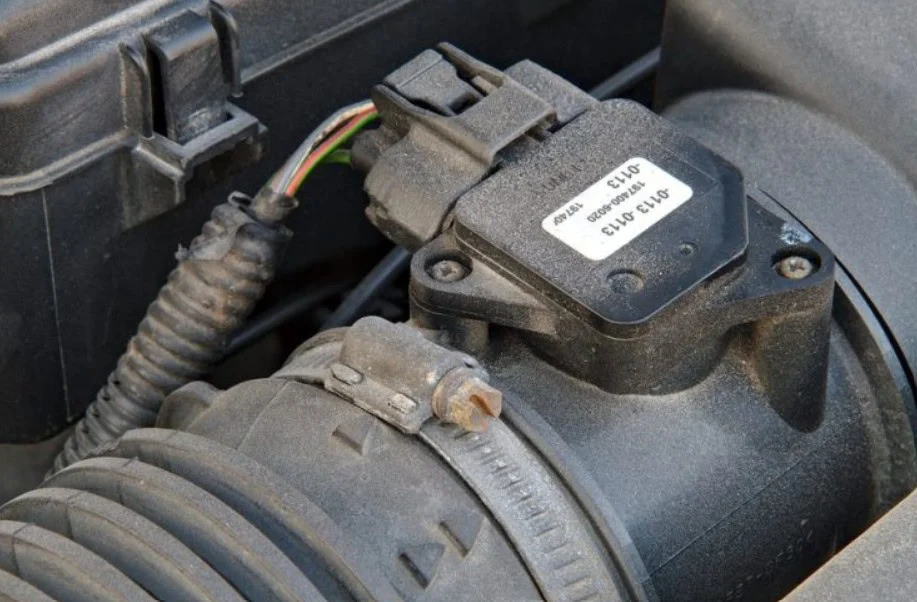Mass air flow sensor with electrical connector and identification label installed on an automotive intake hose near the engine air filter.