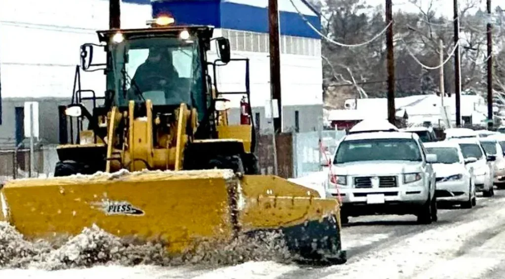 A yellow snowplow clearing snow on a road with a line of cars behind it, performing routine engine air filter maintenance.