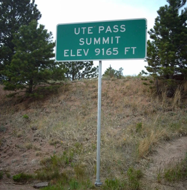 Road sign on a metal pole in a wooded area marking Ute Pass Summit at an elevation of 9165 feet, a point for high-altitude driving.