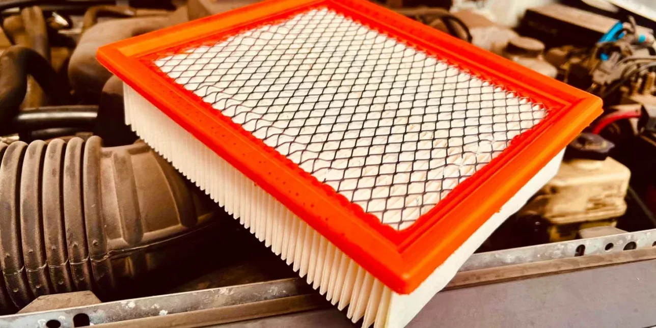 Rectangular engine air filter with a pleated white paper element and an orange plastic frame, resting on an open vehicle hood on Yellowstone Avenue in Pocatello, Idaho.