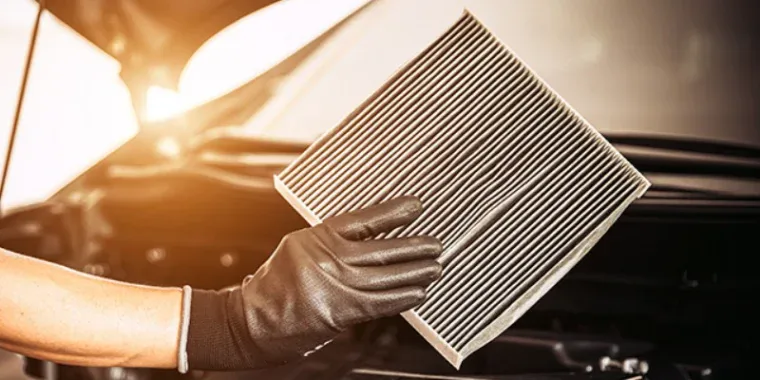 A person wearing a black glove holds a rectangular engine air filter in front of an open car hood during routine car maintenance.