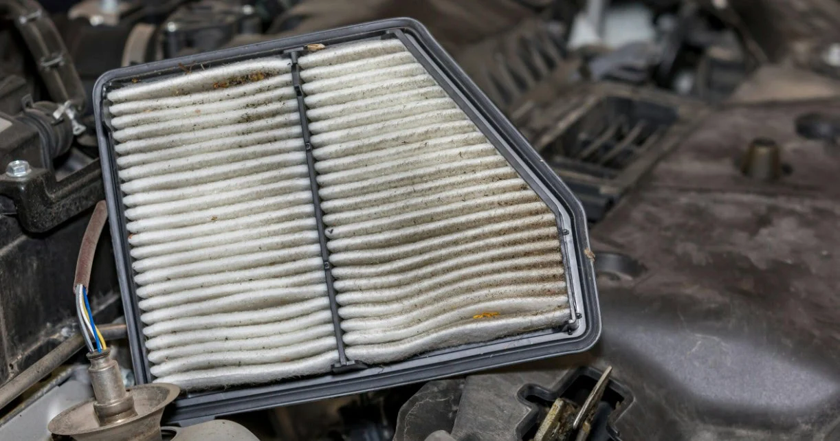 A dirty rectangular engine air filter held above an open car engine bay during a truck air intake inspection.