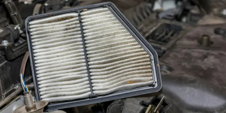 A dirty rectangular engine air filter held above an open car engine bay during a truck air intake inspection.