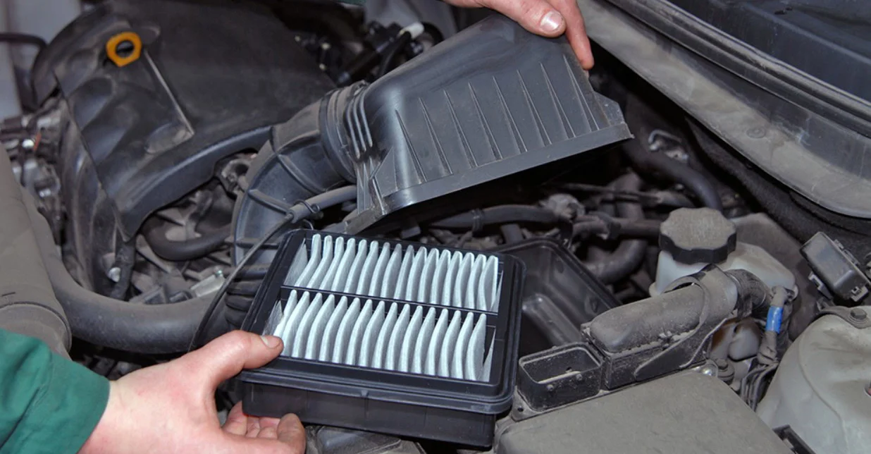 A person lifts the cover of a car’s engine air filter housing to replace the rectangular pleated air filter during routine car maintenance.