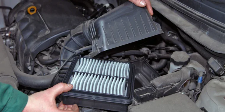 A person lifts the cover of a car’s engine air filter housing to replace the rectangular pleated air filter during routine car maintenance.