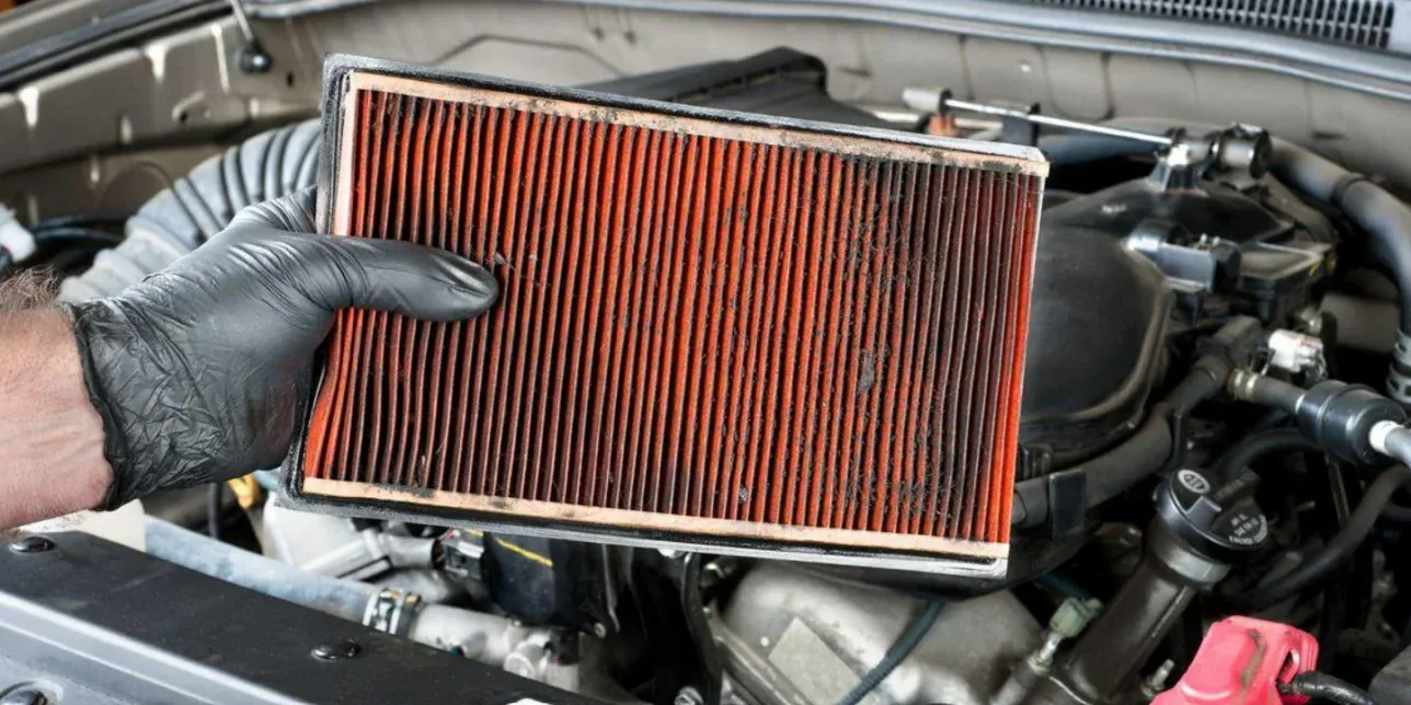 A person wearing a black glove holds a dirty, used engine air filter during farm vehicle maintenance.