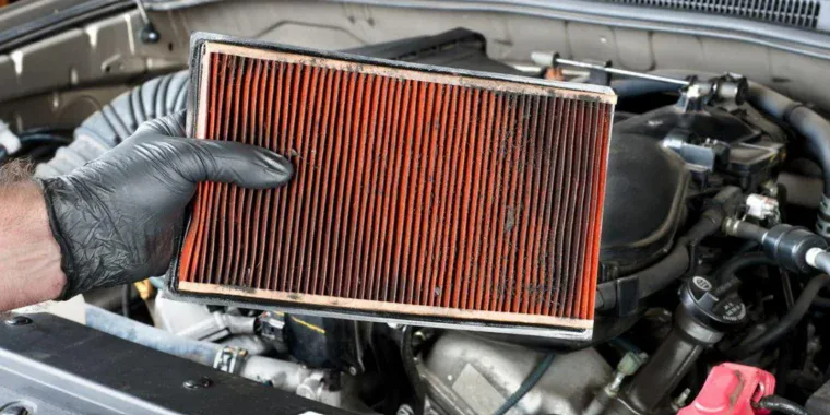 A person wearing a black glove holds a dirty, used engine air filter during farm vehicle maintenance.