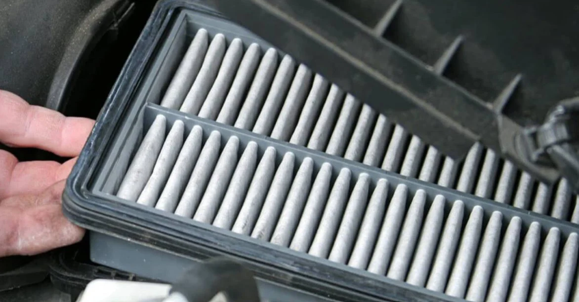 A hand holds open a car’s engine air filter housing to reveal a pleated rectangular filter during routine maintenance.