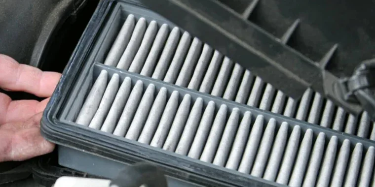 A hand holds open a car’s engine air filter housing to reveal a pleated rectangular filter during routine maintenance.