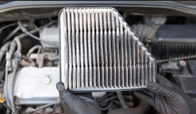 Hands wearing black gloves hold a dirty, dust-covered engine air filter during Aurora car maintenance.